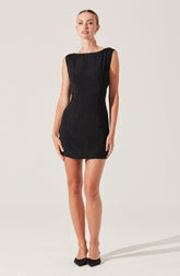 A woman is wearing a sleeveless, fitted black dress with a textured fabric pattern. The dress has a boat neckline and falls to mid-thigh length. She is also wearing black shoes with a bow detail on the front.